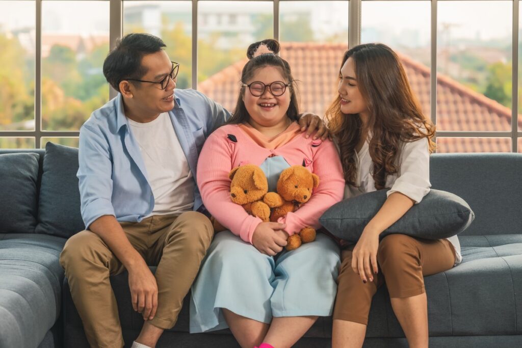 Adolescent girl with intellectual disability sits between her parents smiling and holding two teddy bears.