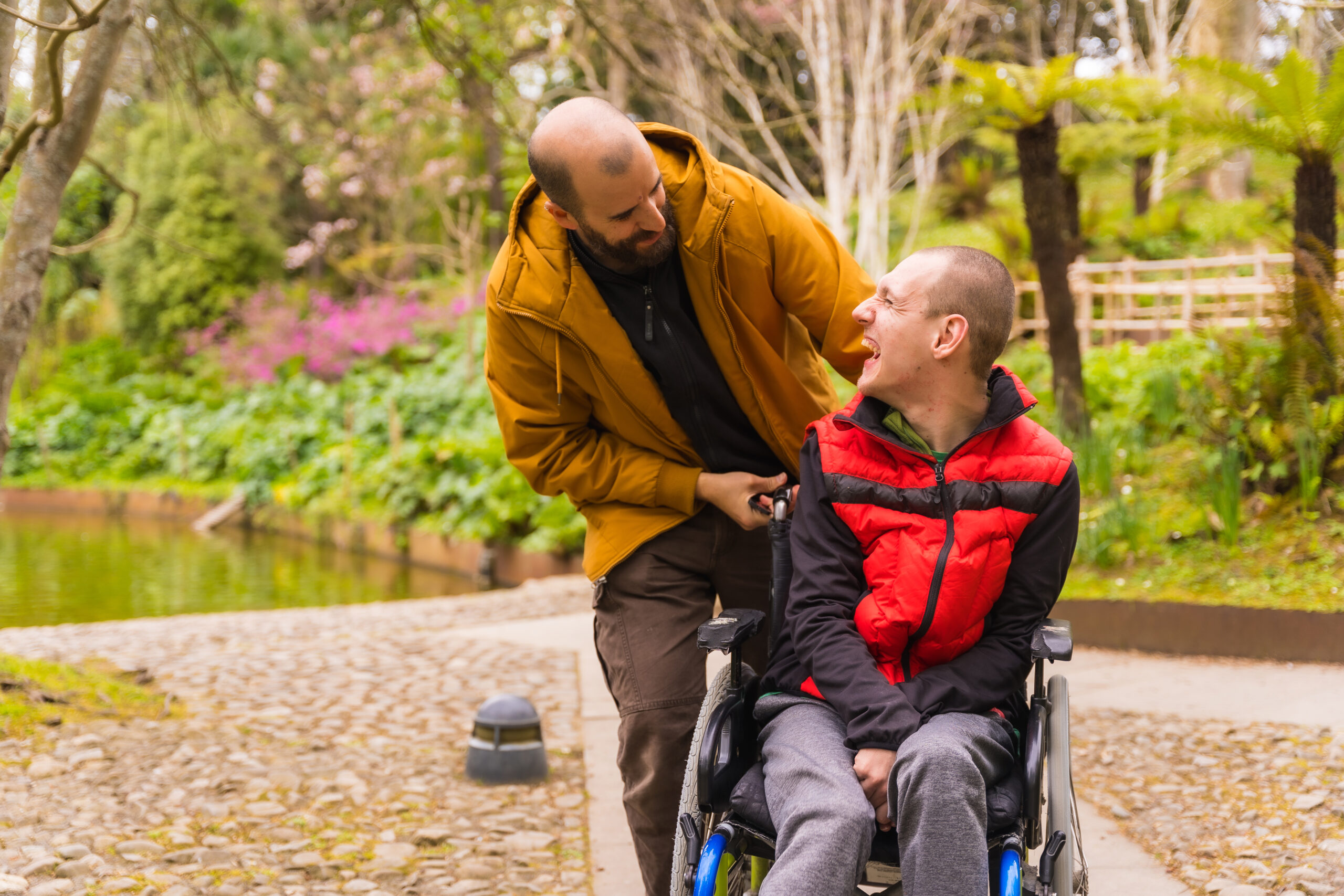Young man in wheelchair having fun being pushed by family caregiver in city park.